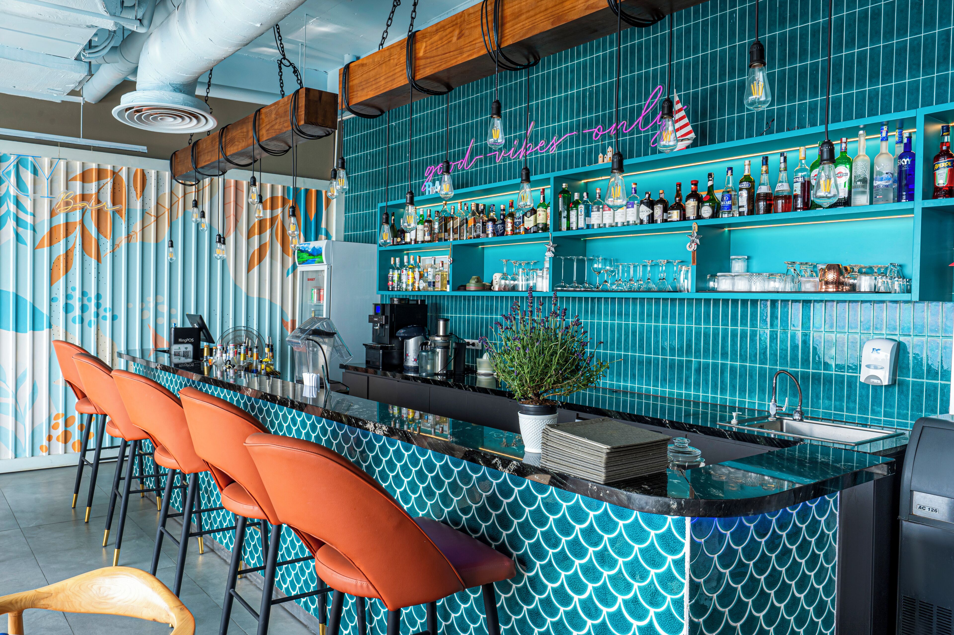 Una zona de bar con un revestimiento de azulejos color turquesa, taburetes naranjas y una variedad de botellas y vasos en estantes.
