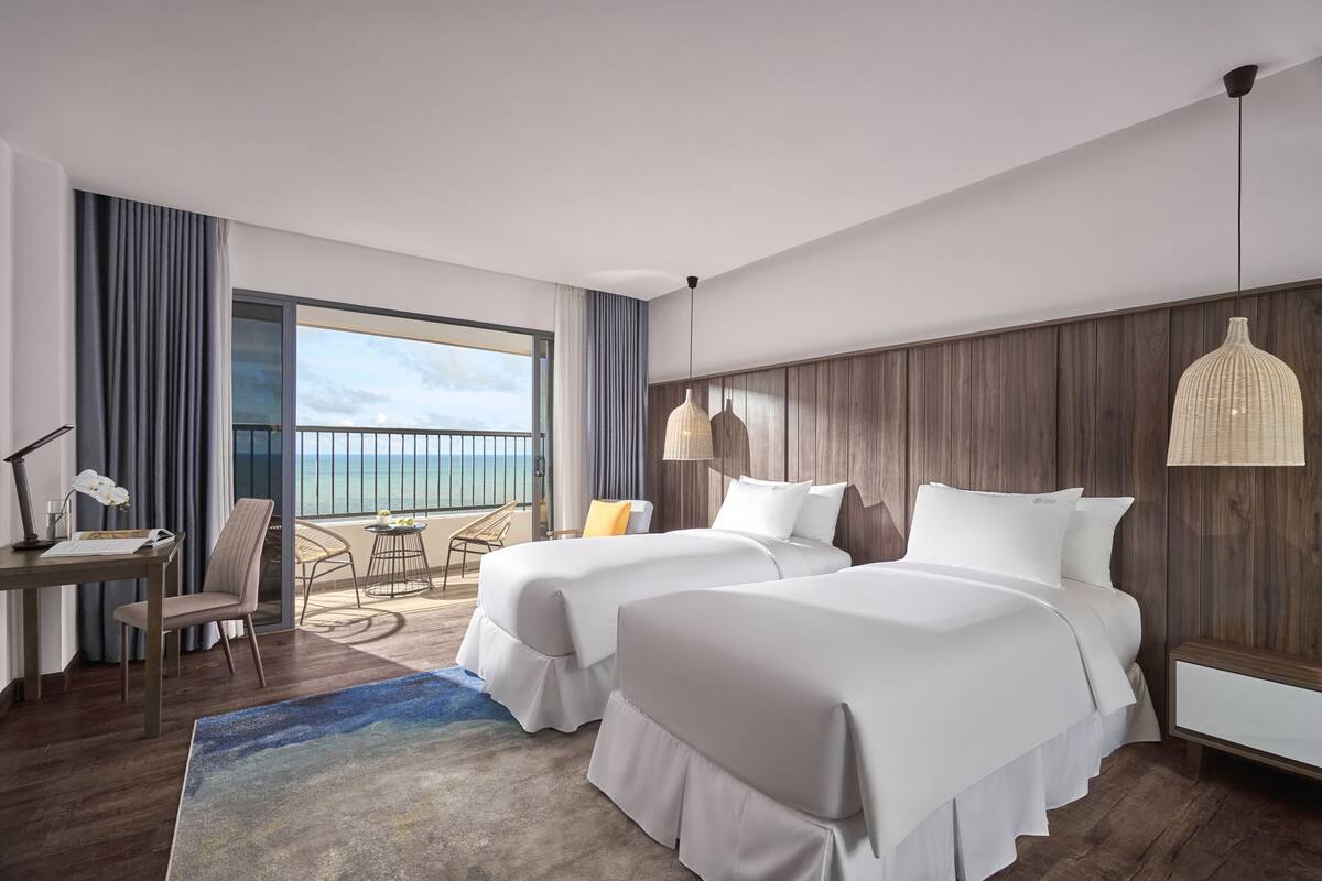 A hotel room with two beds, a desk, a chair, a lamp, and a view of the ocean.