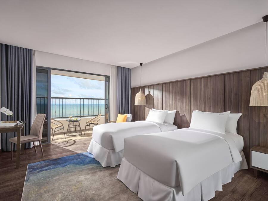 A hotel room with two beds, a desk, a chair, a lamp, and a view of the ocean.