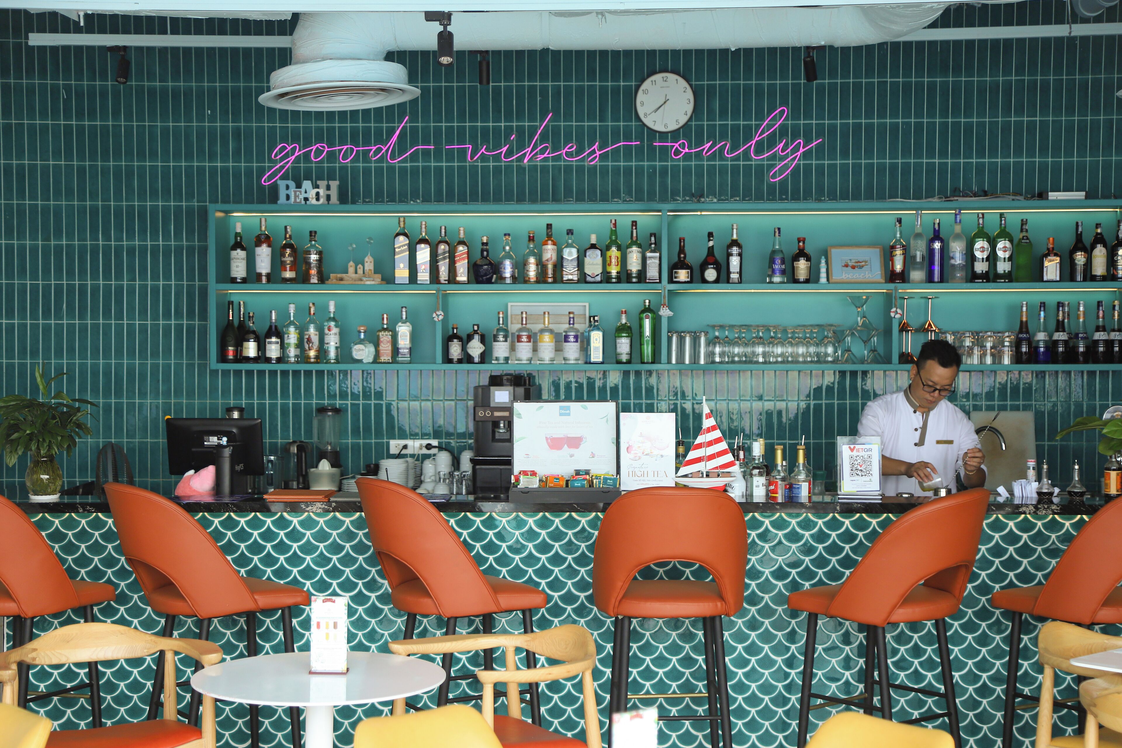 Eine Bar mit einer türkisfarben gefliesten Wand, einem Neon-Schild mit der Aufschrift "good vibes only" und einem Barkeeper, der Getränke zubereitet.
