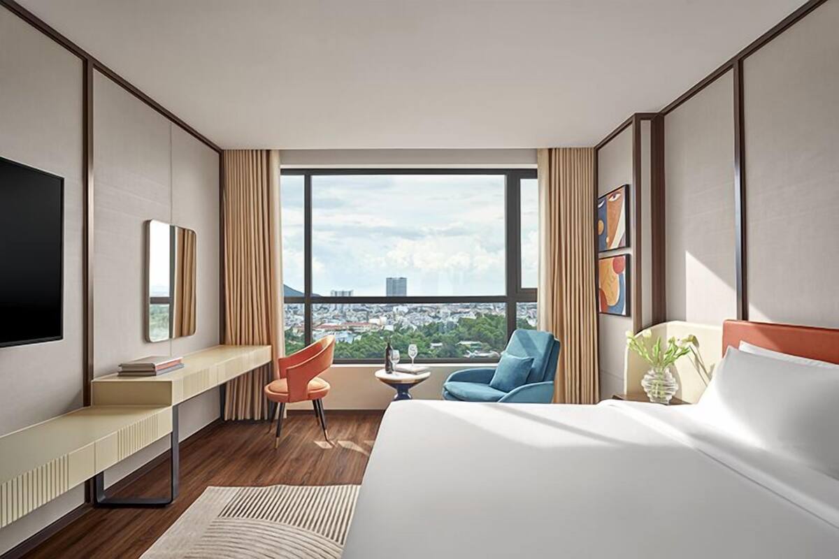 A modern hotel room with a large bed, a desk, a chair, a small table, and a large window offering a city view.