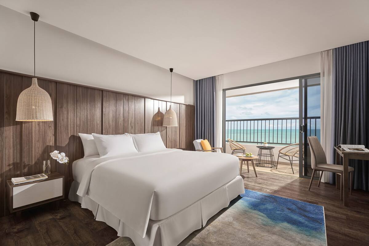 A hotel room with a large bed, a nightstand, a reading lamp, a chair, a small table, and a view of the ocean through a balcony.