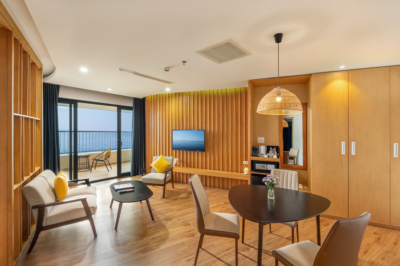 A modern hotel room with a balcony, wooden panel walls, a dining area, and a TV mounted on the wall.
