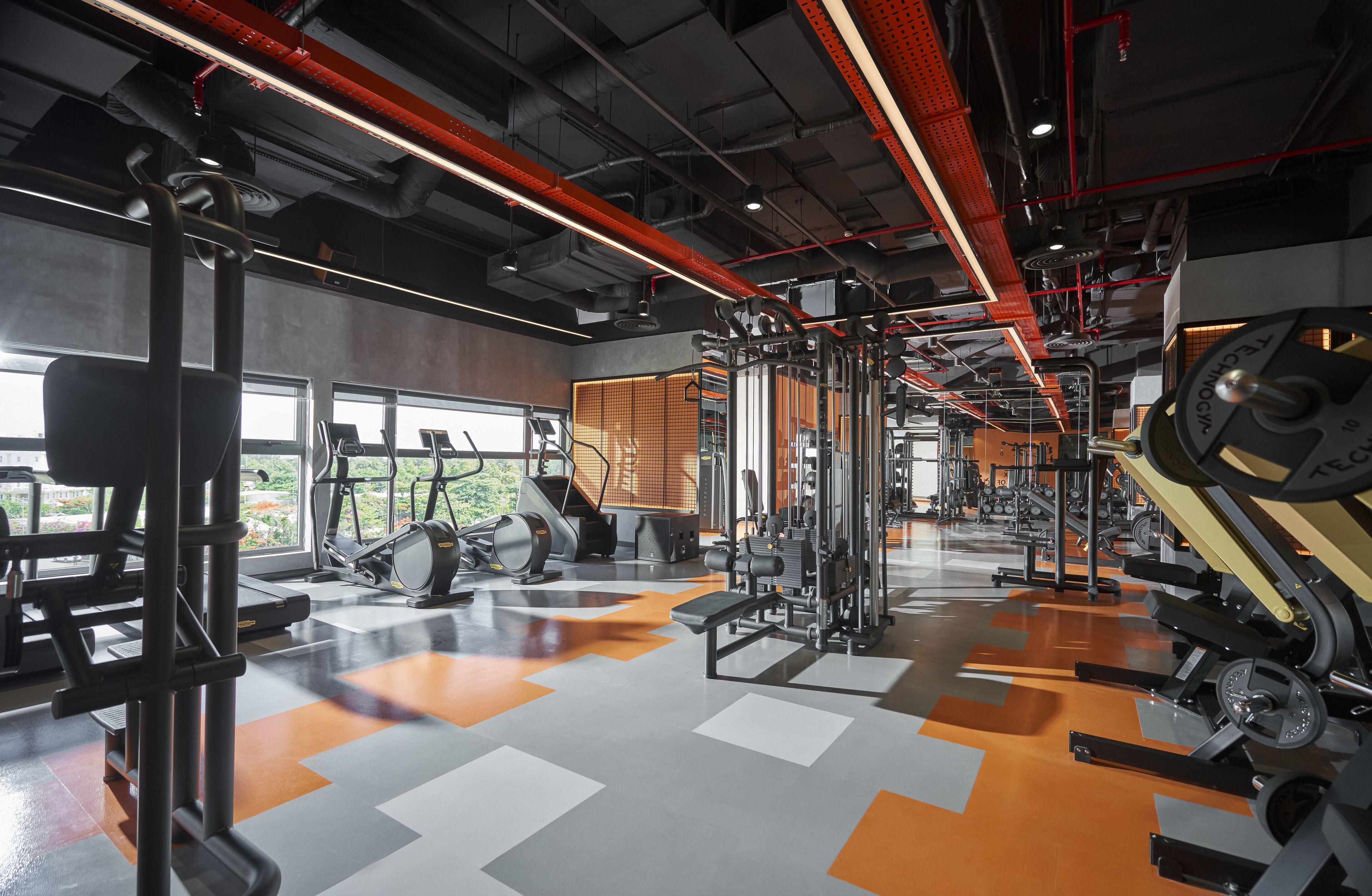 Une salle de sport équipée de divers appareils d’exercice, dotée de grandes fenêtres et d’un sol à motifs en damier.