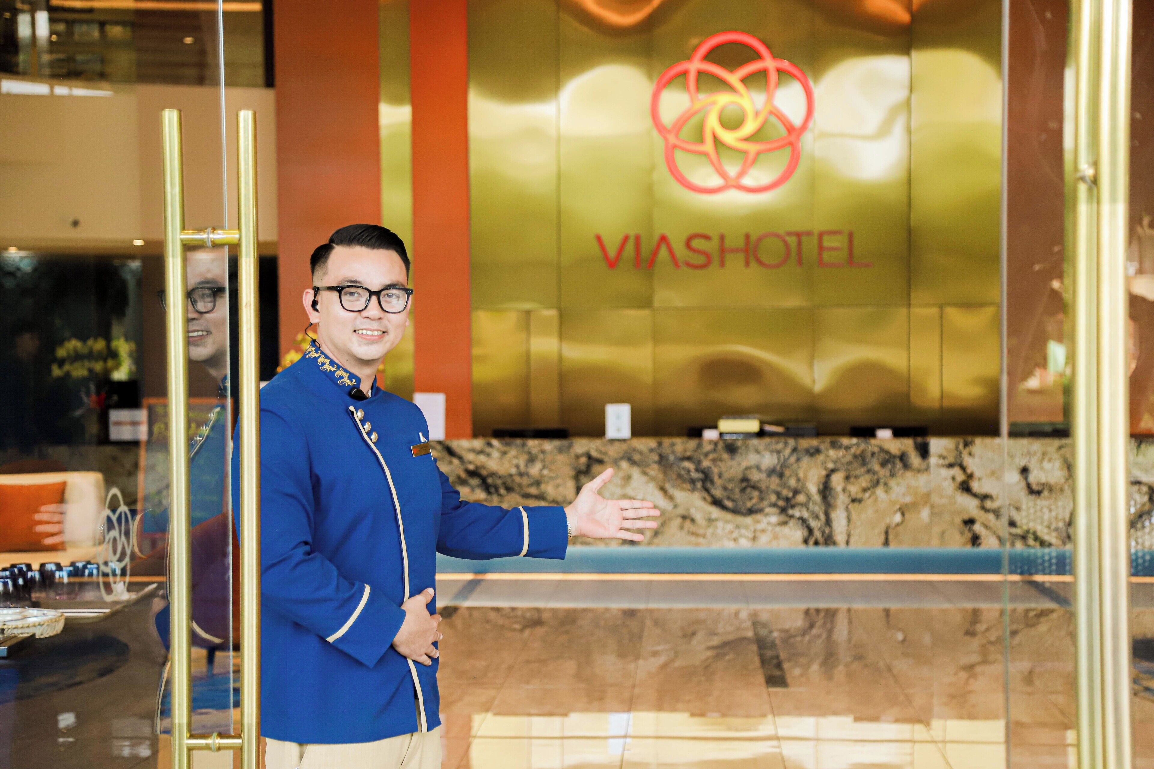 Une personne se tient devant un comptoir d’accueil, sur le mur duquel figure un logo et le nom « VIASHOTEL ».