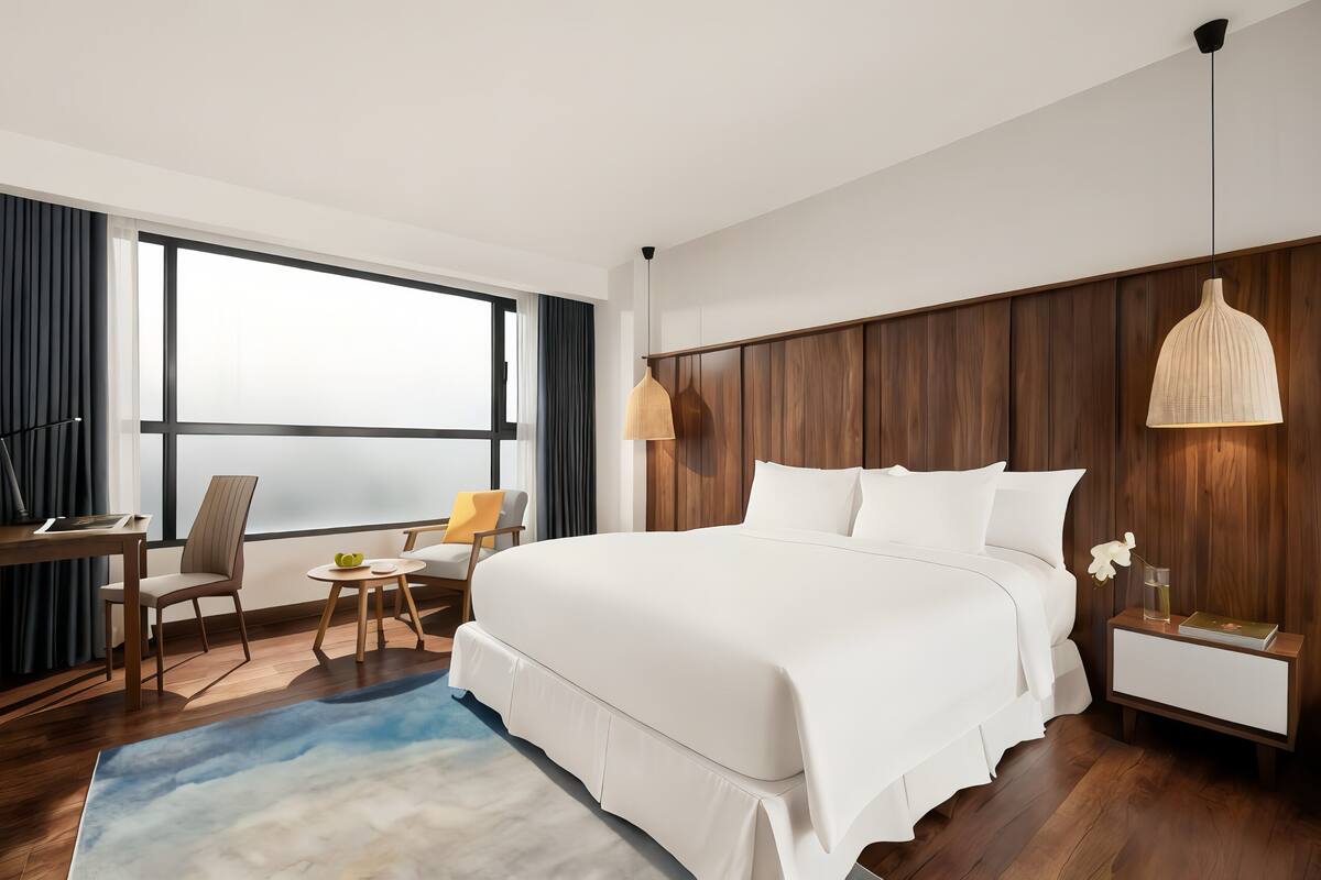 A hotel room with a large bed, a desk, a chair, a small table, a nightstand, and a large window.