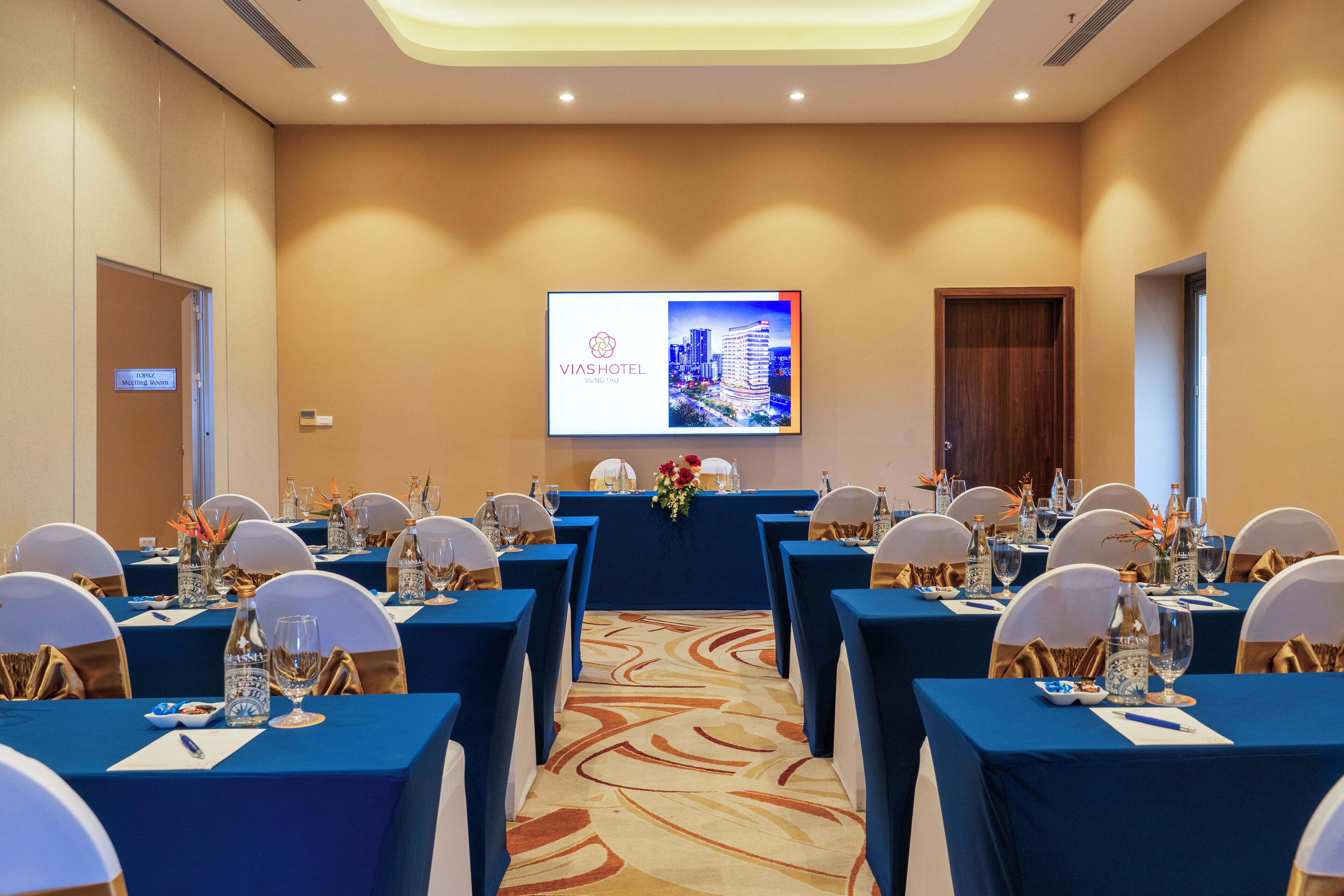 A conference room with tables set for a meeting, a presentation screen, and a door labeled 'TOPAZ Meeting Room'.