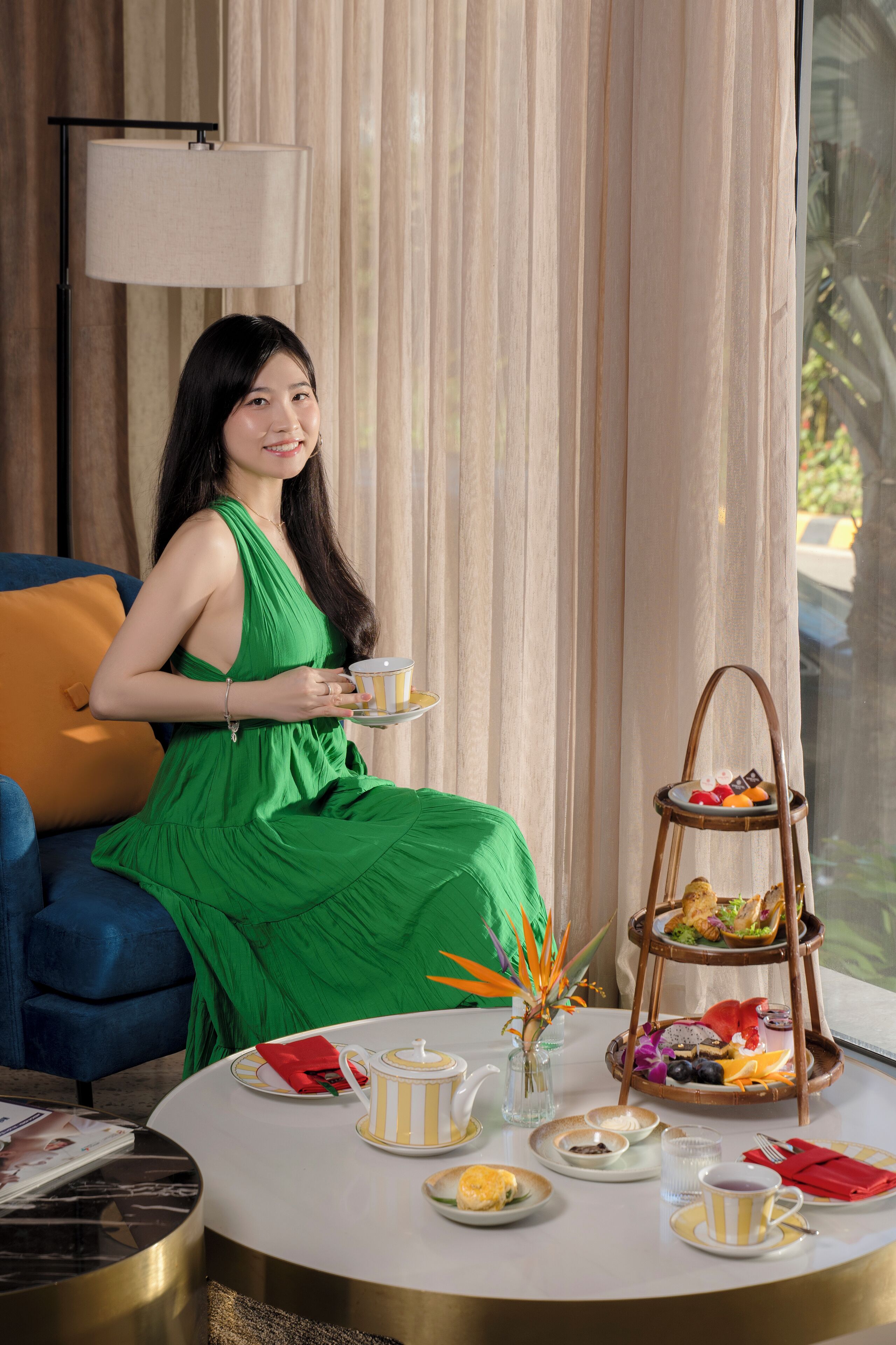 A woman in a green dress sitting on a blue sofa in a room with a round table set for tea, a lamp, and a window with curtains.