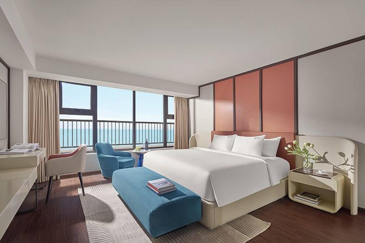 A modern hotel room with a large bed, a desk, a chair, a small table with a vase, and a view of the sea.