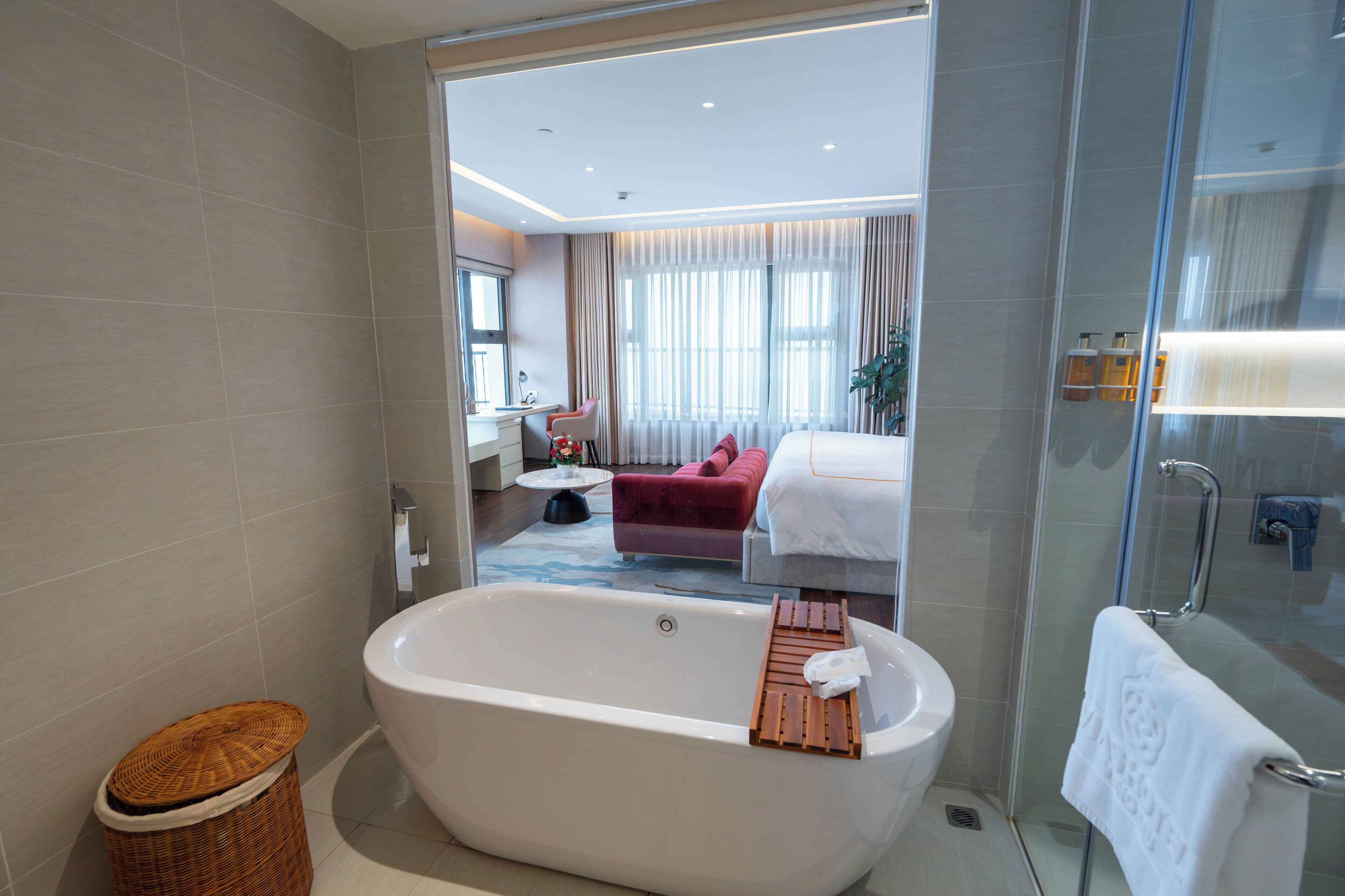 Un baño moderno con una bañera independiente, cabina de ducha con mampara de vidrio, y se ve un dormitorio con un sofá rojo y una cama.