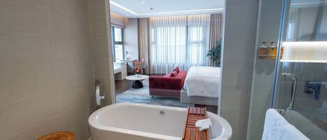 Un baño moderno con una bañera independiente, cabina de ducha con mampara de vidrio, y se ve un dormitorio con un sofá rojo y una cama.