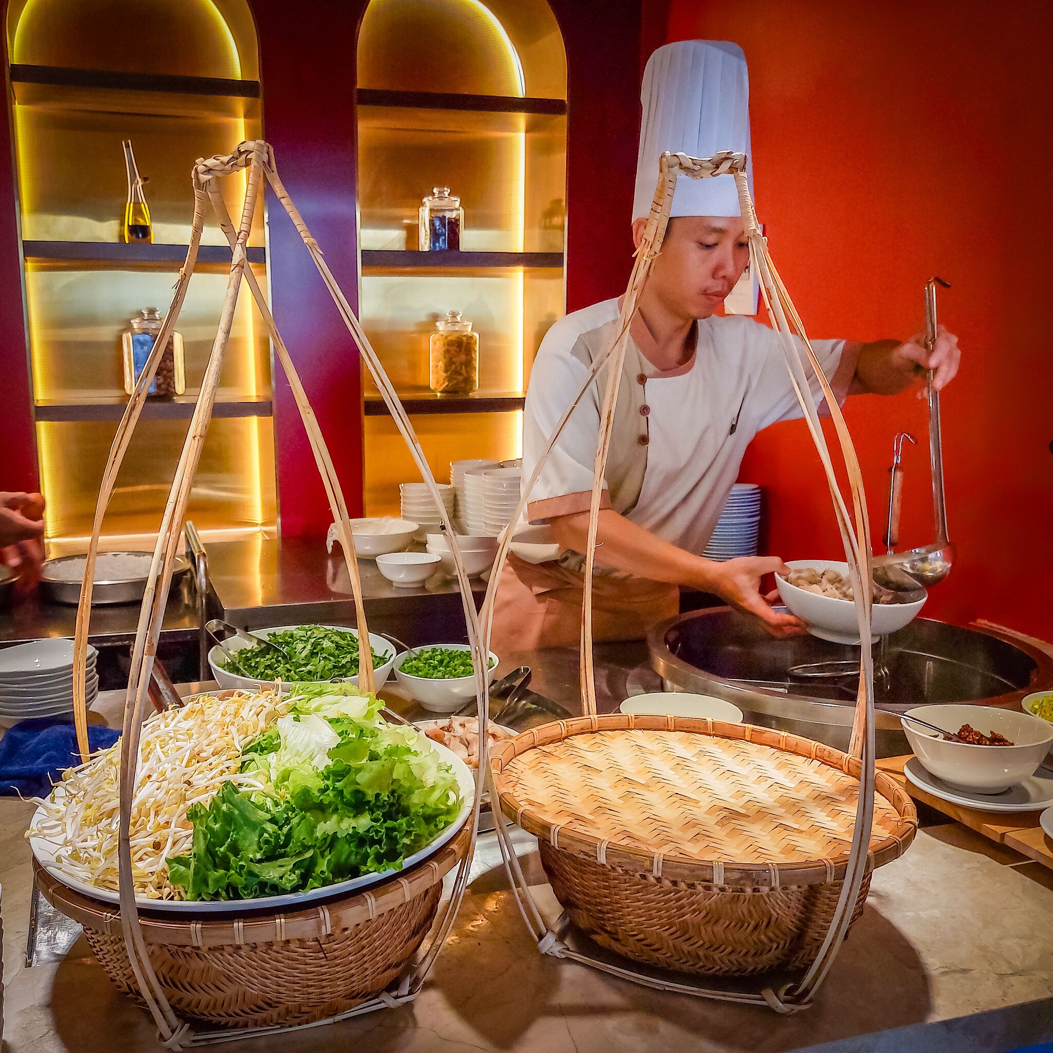 Een kok met een traditionele hoed bereidt eten in een keuken vol ingrediënten en keukengerei.