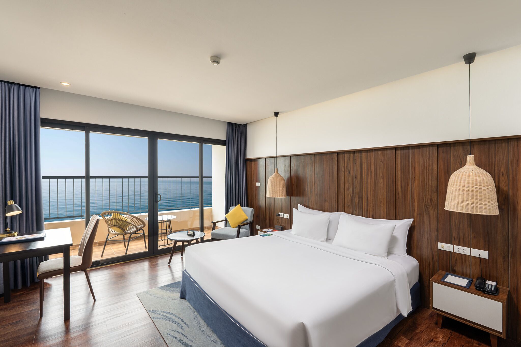 Habitación de hotel moderna con una cama grande, un escritorio y una silla. Se ve el océano a través de amplios ventanales.