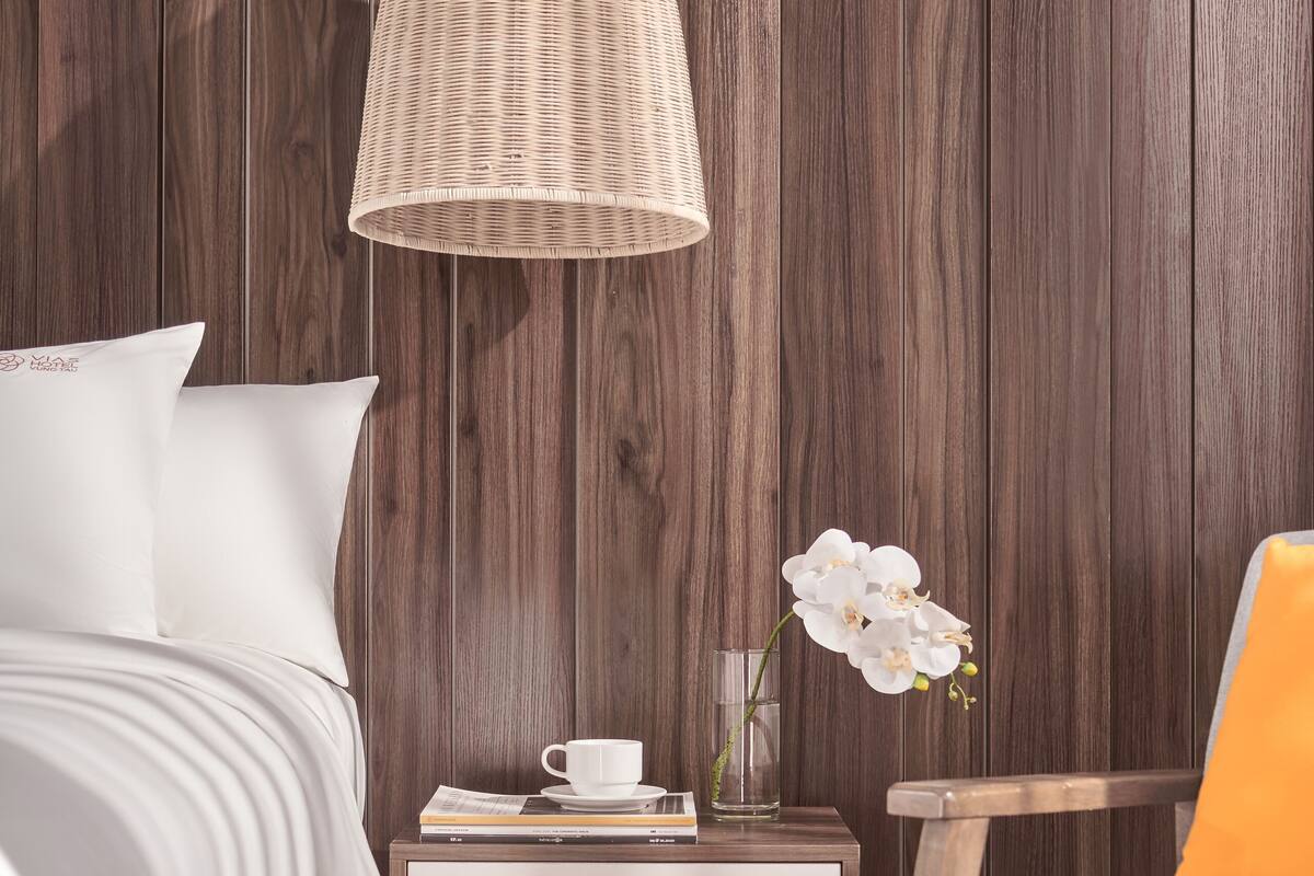 A hotel room with a wooden panel wall, a bed with white bedding, a nightstand with a cup and book, a chair with an orange cushion, and a hanging light fixture.