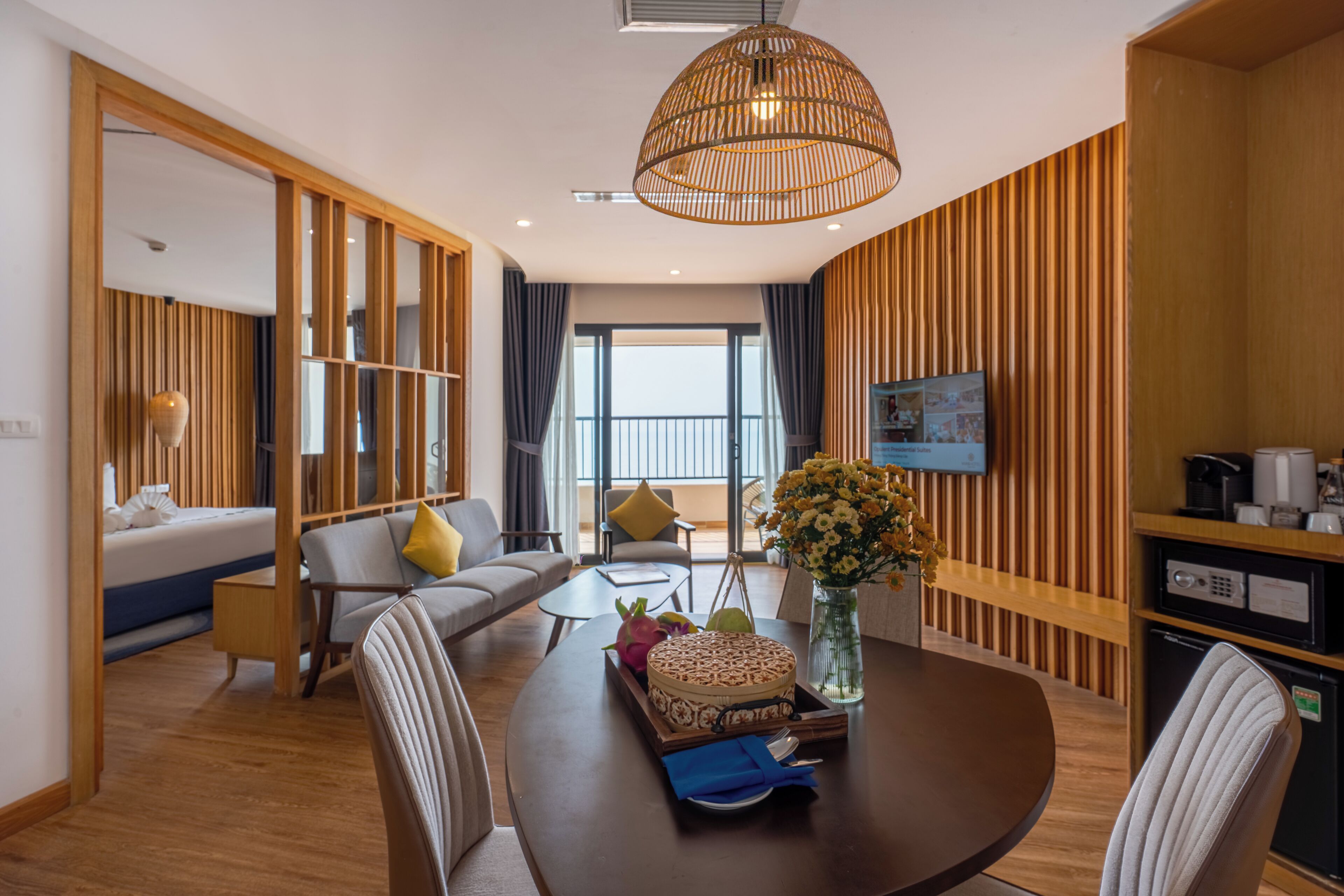 A modern hotel room with a large bed, a sofa, a dining table with a vase of flowers, and a television on the wall.