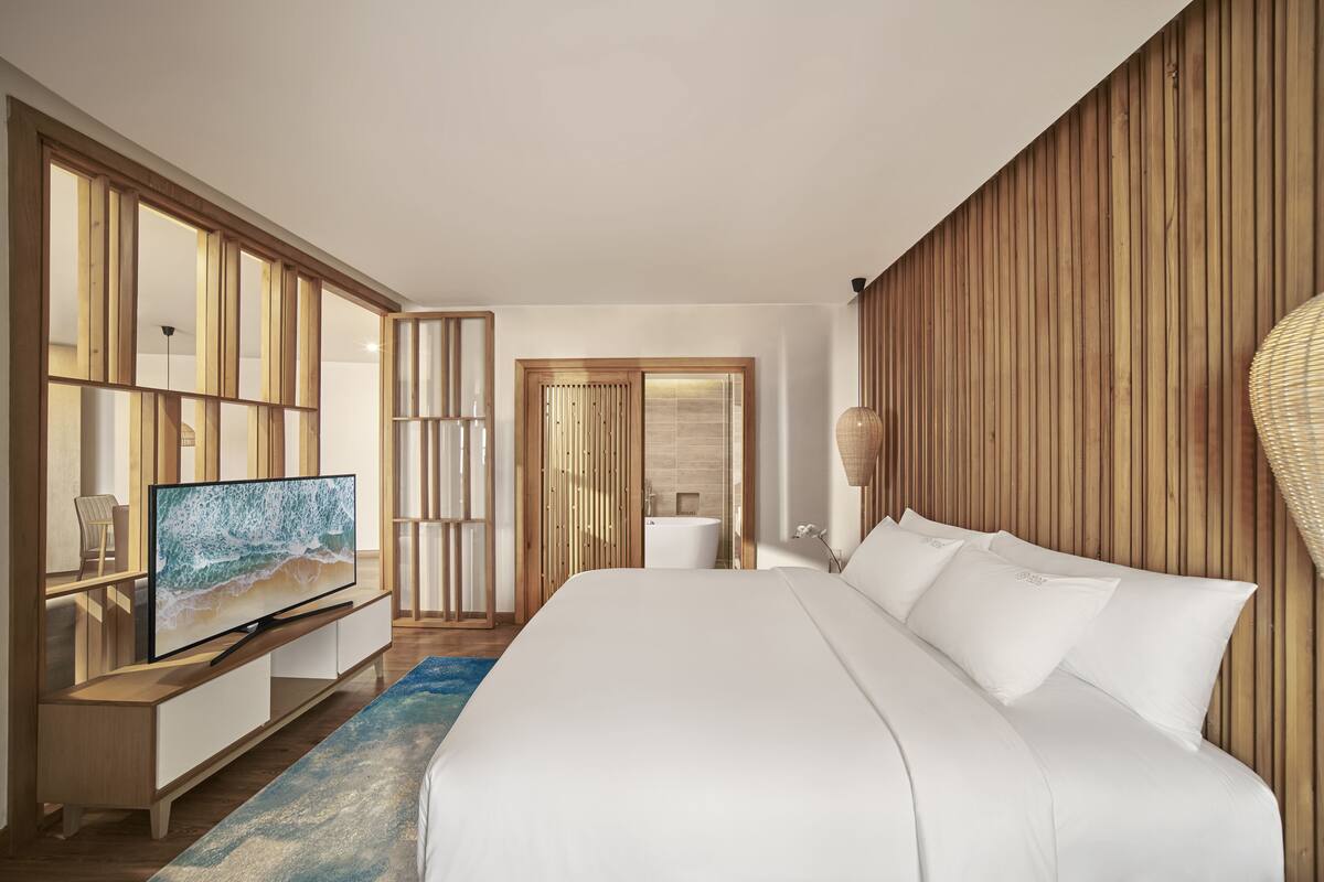 A modern hotel room with a large bed, a flat-screen TV, a wooden accent wall, and a bathroom visible through an open door.