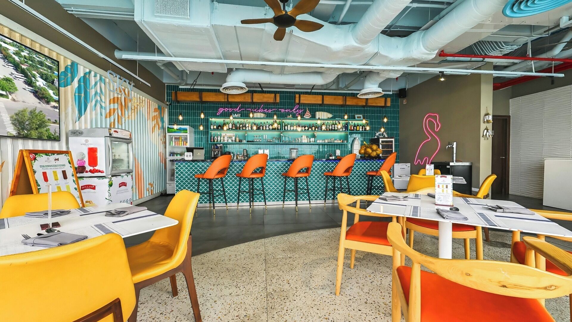 A bar area with a counter, bar stools, and a neon sign. There are tables with chairs and a ceiling fan.