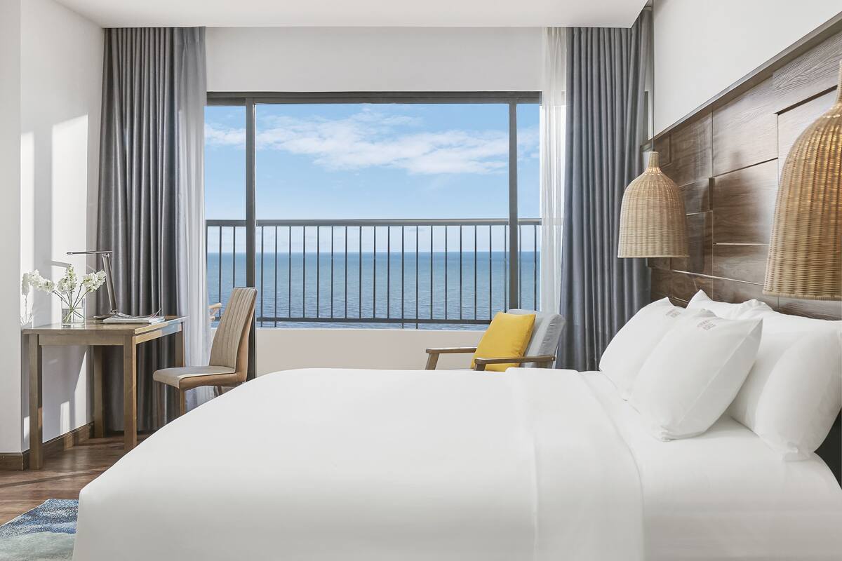 A hotel room with a large bed, a desk with a lamp, a chair, and a view of the sea through a balcony.