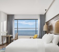 Hotellihuone, jossa on suuri sänky, työpöytä lampun kanssa, tuoli ja parvekkeelta avautuva merinäköala.