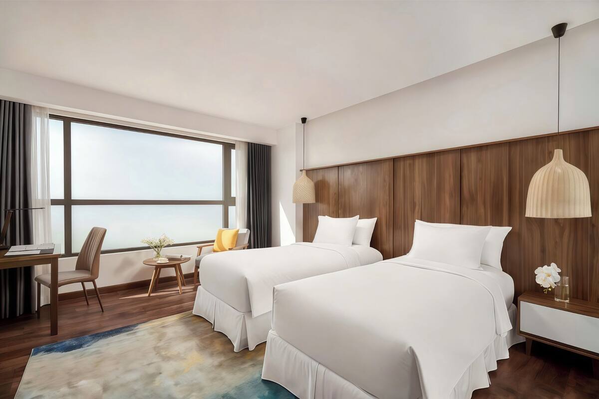 A hotel room with two beds, a desk, a chair, a small table with a vase, and a large window with curtains.