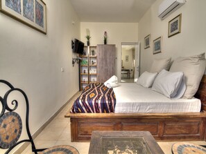 Quarto com cama de madeira, televisão na parede, mesinha e prateleira com objetos decorativos.