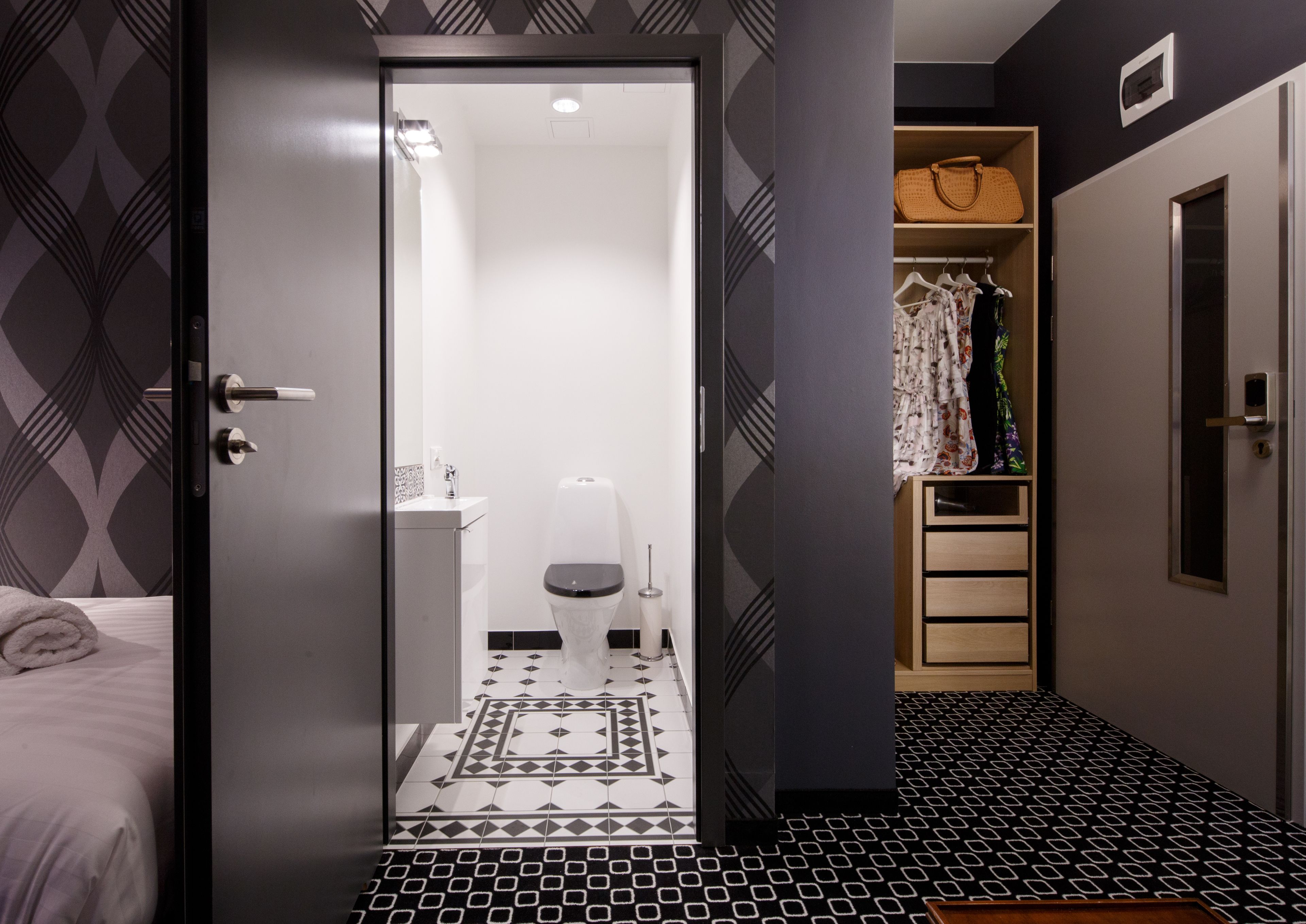 A modern hotel room with a geometric patterned floor, a white toilet, a wooden wardrobe, and a bed with a rolled towel.