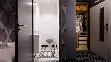 A modern hotel room with a geometric patterned floor, a white toilet, a wooden wardrobe, and a bed with a rolled towel.