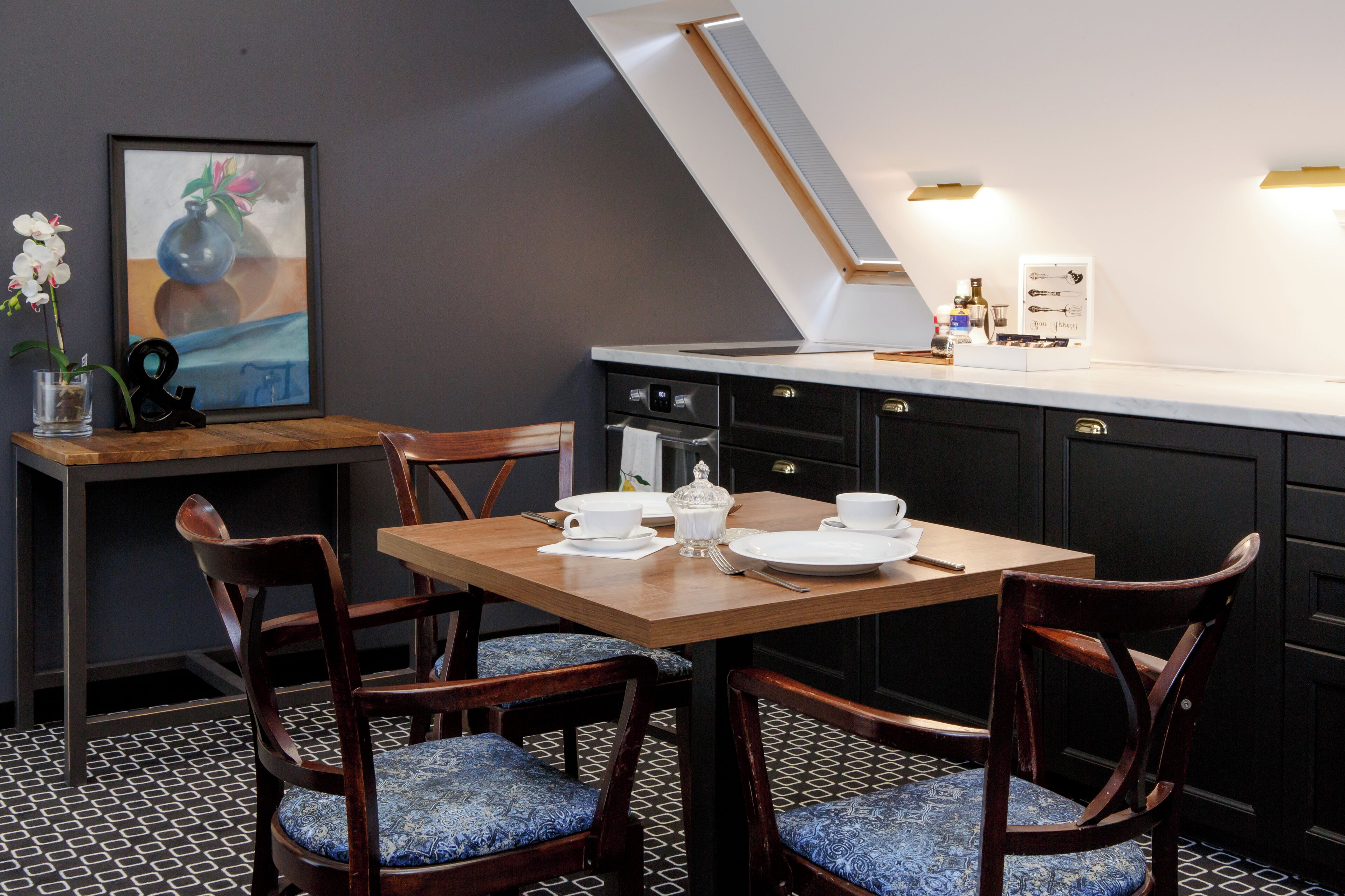 A dining area with a wooden table, chairs with patterned cushions, a framed artwork, and a kitchen counter with appliances.