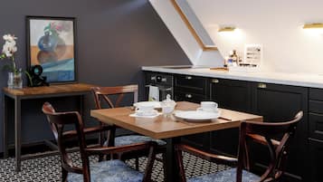A dining area with a wooden table, chairs with patterned cushions, a framed artwork, and a kitchen counter with appliances.