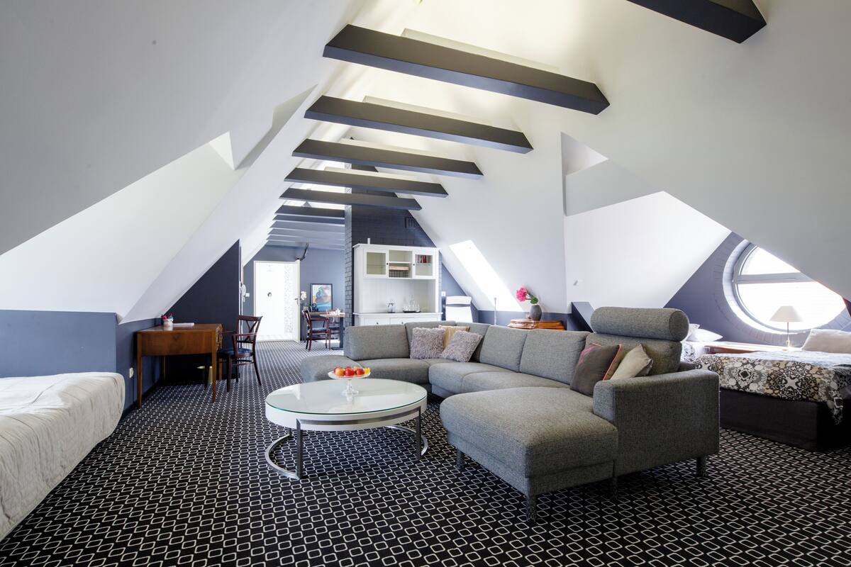 Ein modernes Schlafzimmer im Loft-Stil mit schrägen Wänden, einem grauen Ecksofa, einem runden Couchtisch und einem großen Fenster, das für natürliches Licht sorgt.