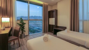 Habitación de hotel con un ventanal grande, un escritorio, una silla, una planta en maceta y dos camas.