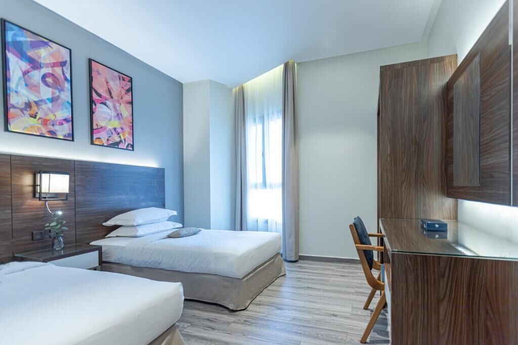 Ein modernes Hotelzimmer mit einem großen Bett, einem Schreibtisch und einem Fernseher. An den Wänden hängen zwei Bilder und es gibt ein Fenster mit Vorhängen.