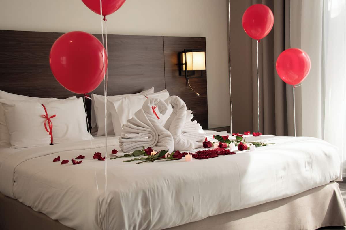 A neatly made bed with white linens, red rose petals, and red balloons.