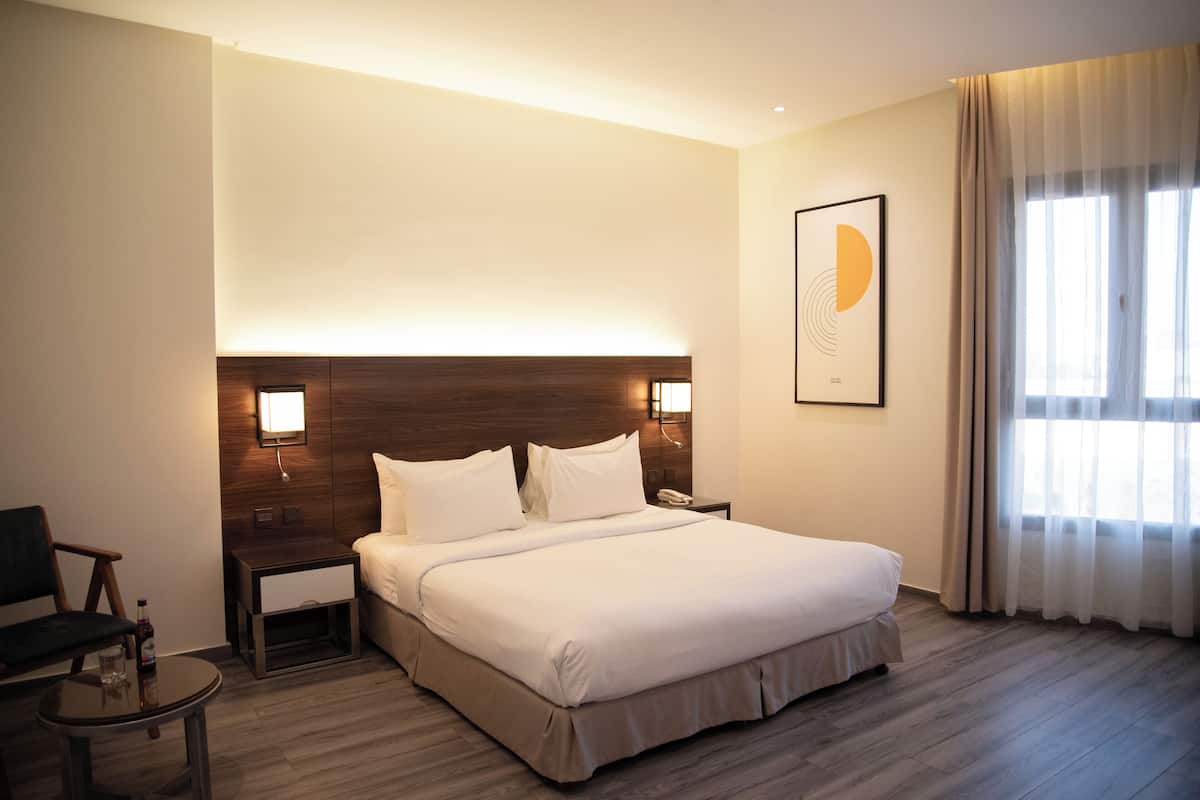 A hotel room with a large bed, a nightstand, a chair, a small table, and a window with curtains.