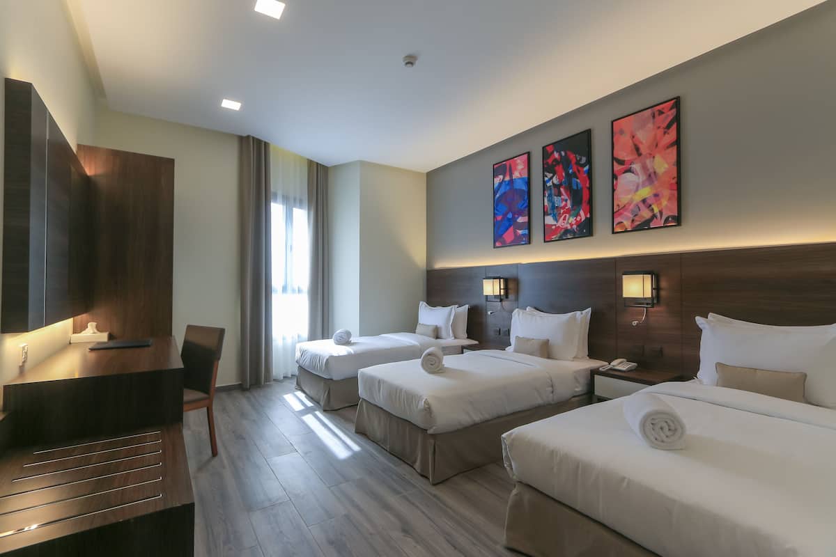 A hotel room with two beds, a desk, and a chair. There are three paintings on the wall and a window with curtains.