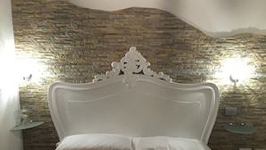 Een slaapkamer met een stenen muur, een wit bed met een decoratief hoofdeinde, en een nachtkastje met een lamp.