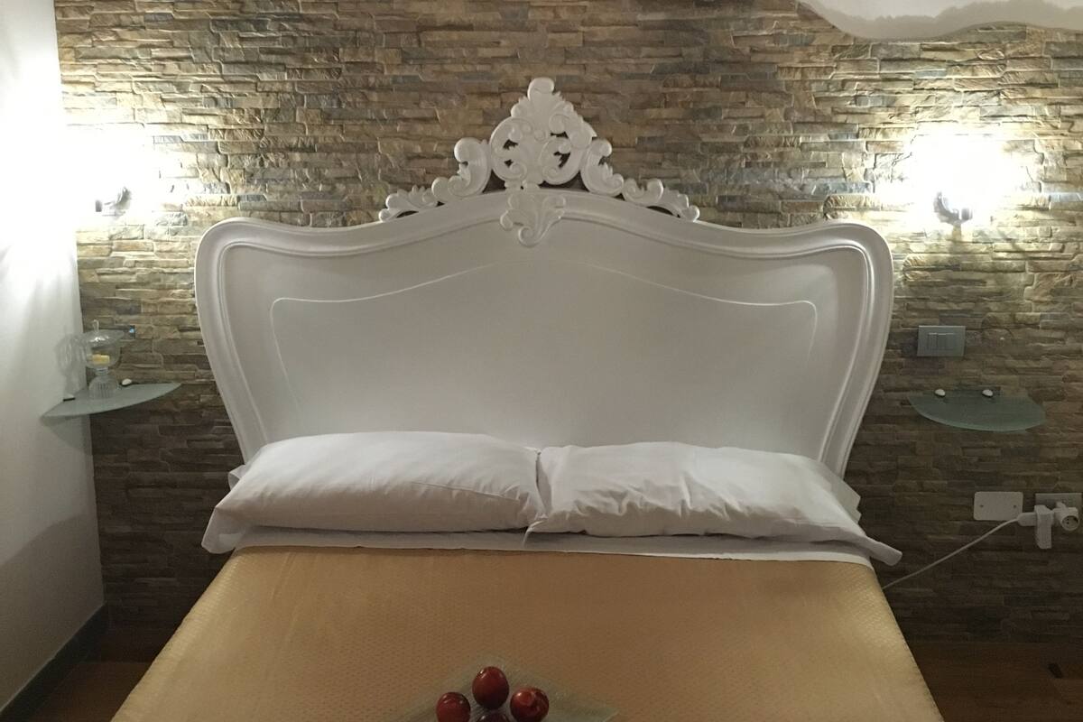 Ein Schlafzimmer mit Steingemäuer, einem weißen Bett mit dekorativer Kopfteil und einem Nachttisch mit Lampe.