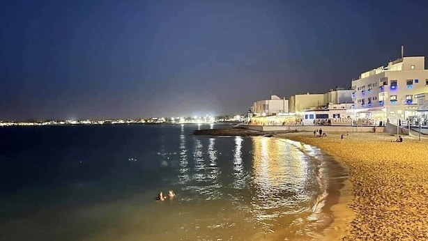En strand i månsken med byggnader längs kusten och några få personer i vattnet.