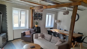 Un salon chaleureux avec un canapé, un fauteuil et une table basse, un coin repas avec une table et des chaises en bois, et une cheminée.