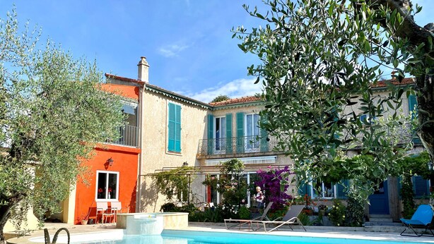 Una piscina con una escultura decorativa, un edificio de fachada roja y un balcón con contraventanas azules.