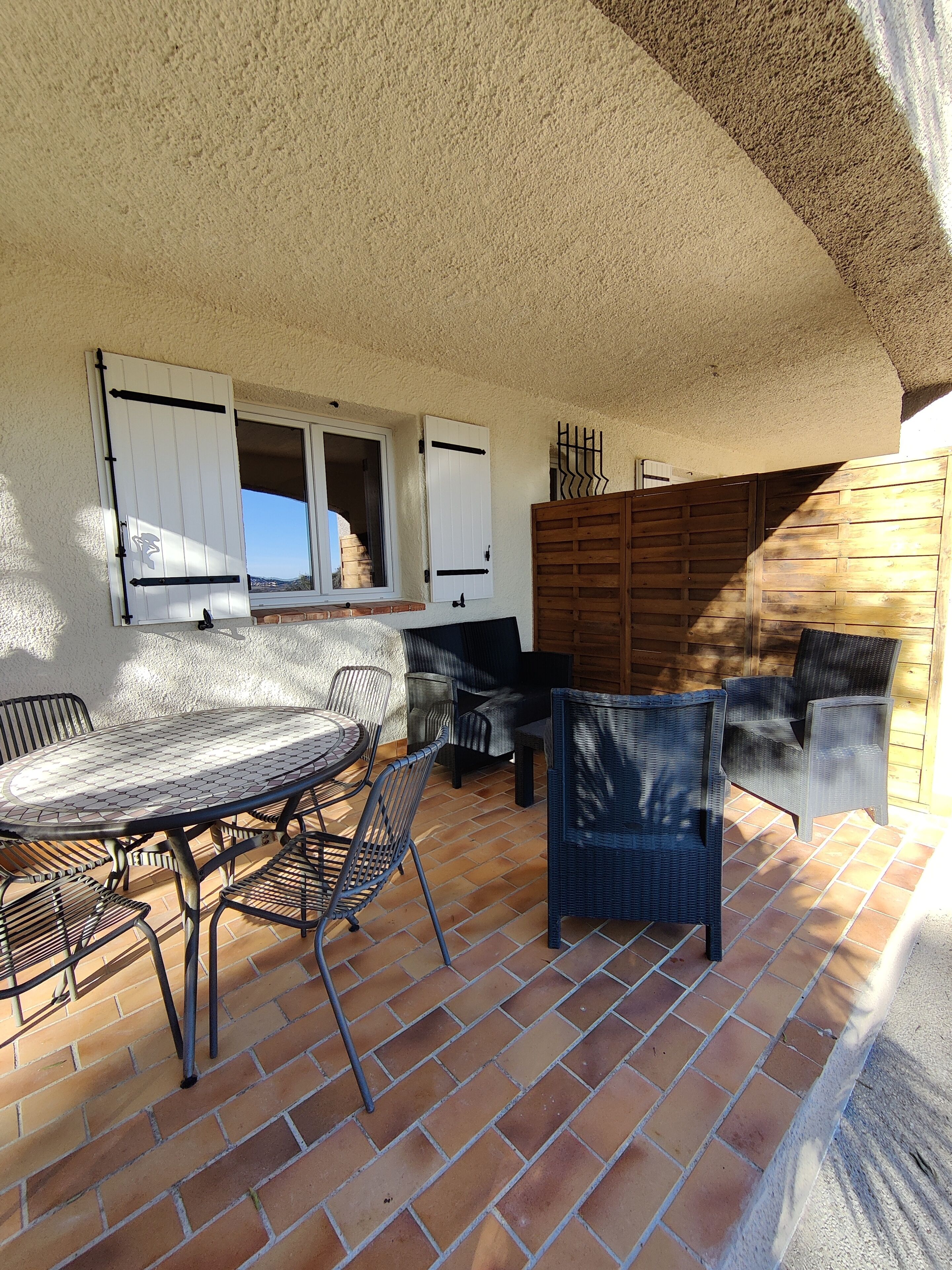 Une terrasse extérieure avec une table et des chaises rondes en métal, une clôture en bois et une fenêtre avec des volets.
