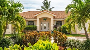 A two-story building with a tiled roof, a central entrance with a decorative arch, and a well-maintained garden with various plants and a palm tree. - P13 - Loggerhead House 3 Bedroom Home with Private Pool (Key Colony Beach)