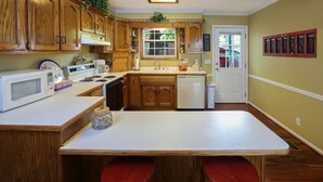 Cocina con armarios de madera, encimera blanca, microondas, estufa, pileta y lavavajillas. Hay una ventana con vista al exterior y una puerta con un cartel arriba.