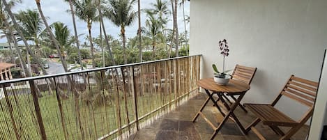 Un balcón con una mesa y sillas de madera, una orquídea en maceta y vistas a palmeras y una calle.