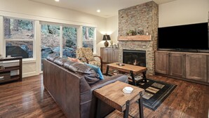 A modern living room with a stone fireplace, a large TV, and a leather sofa. - Trails End Retreat - Perfect Retreat Luxury Home, Guest Quarters, A/C, Foosball, Ping Pong (Bend)