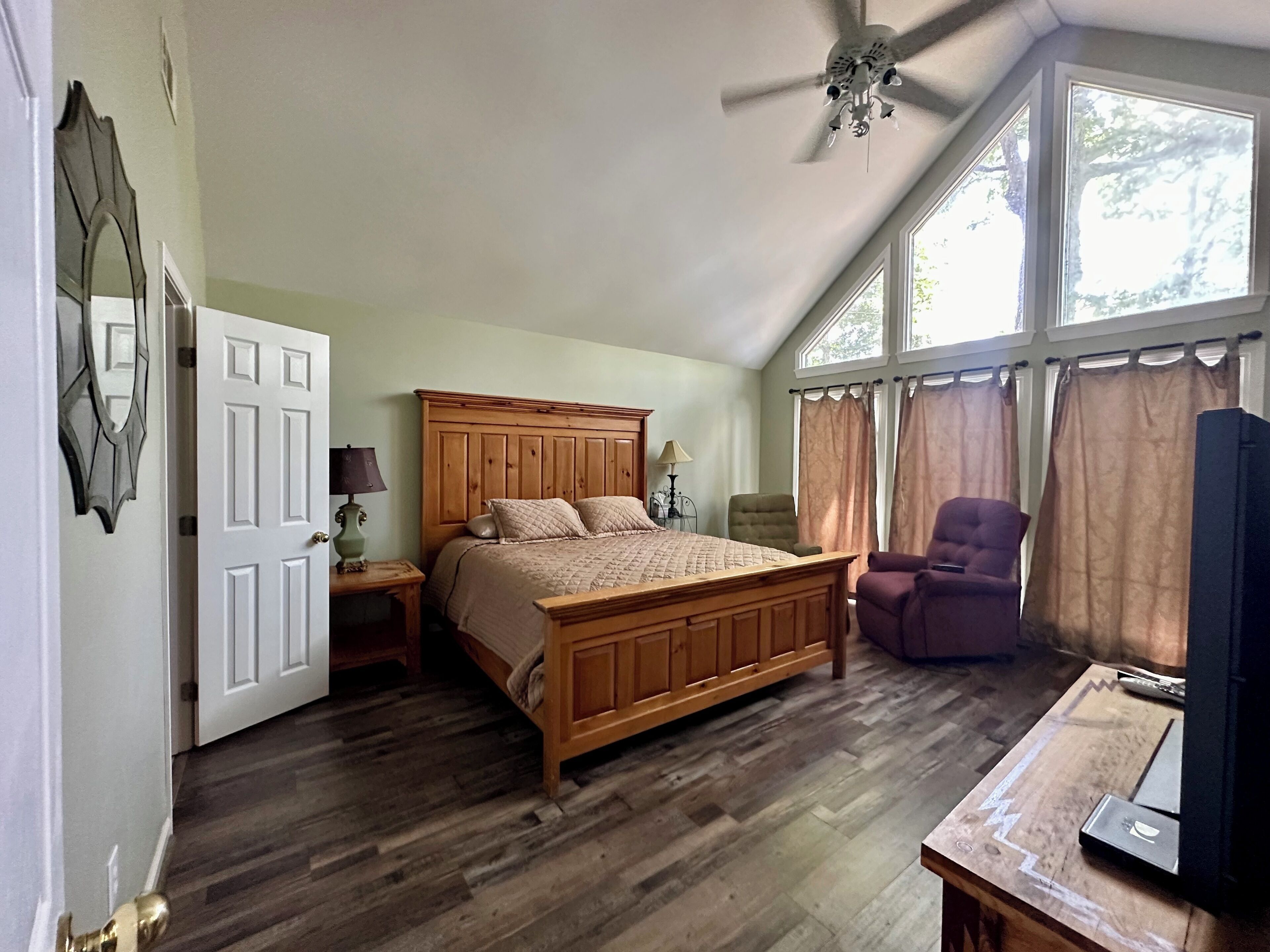 Une chambre avec un lit en bois, un ventilateur de plafond, une chaise, un téléviseur et de grandes fenêtres.