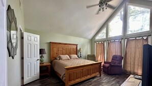 A bedroom with a wooden bed, a ceiling fan, a chair, a TV, and large windows. - Family house by the lake (Shelby)