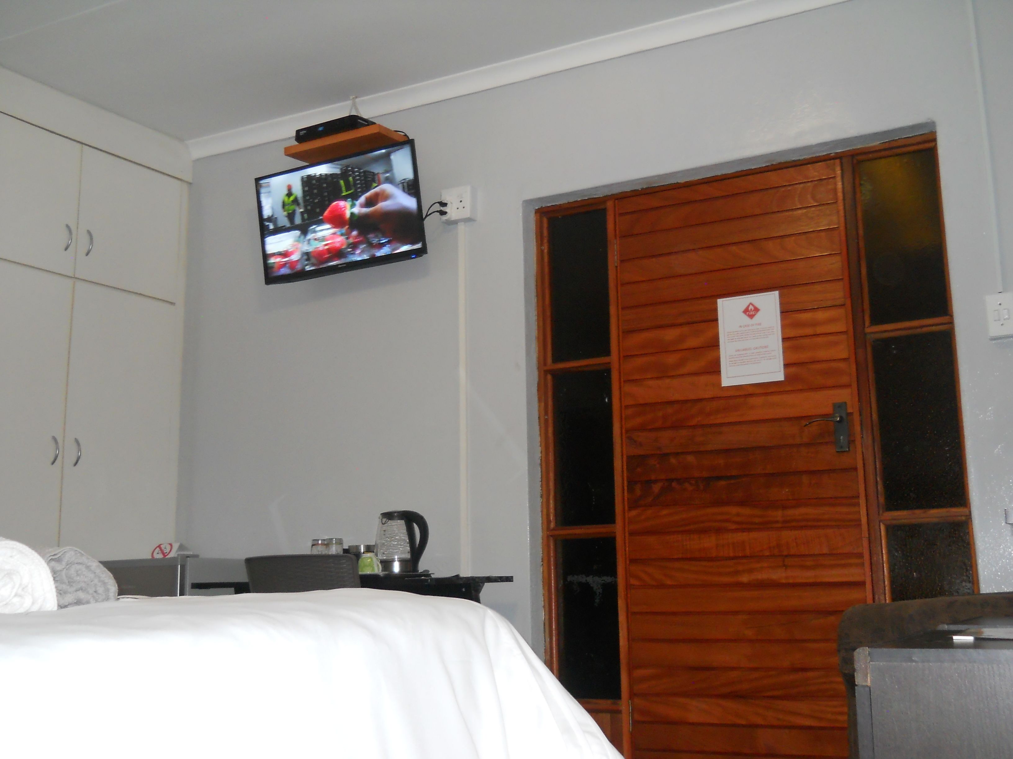 A hotel room with a bed, a television mounted on the wall, a wooden door, and a small table with a kettle.
