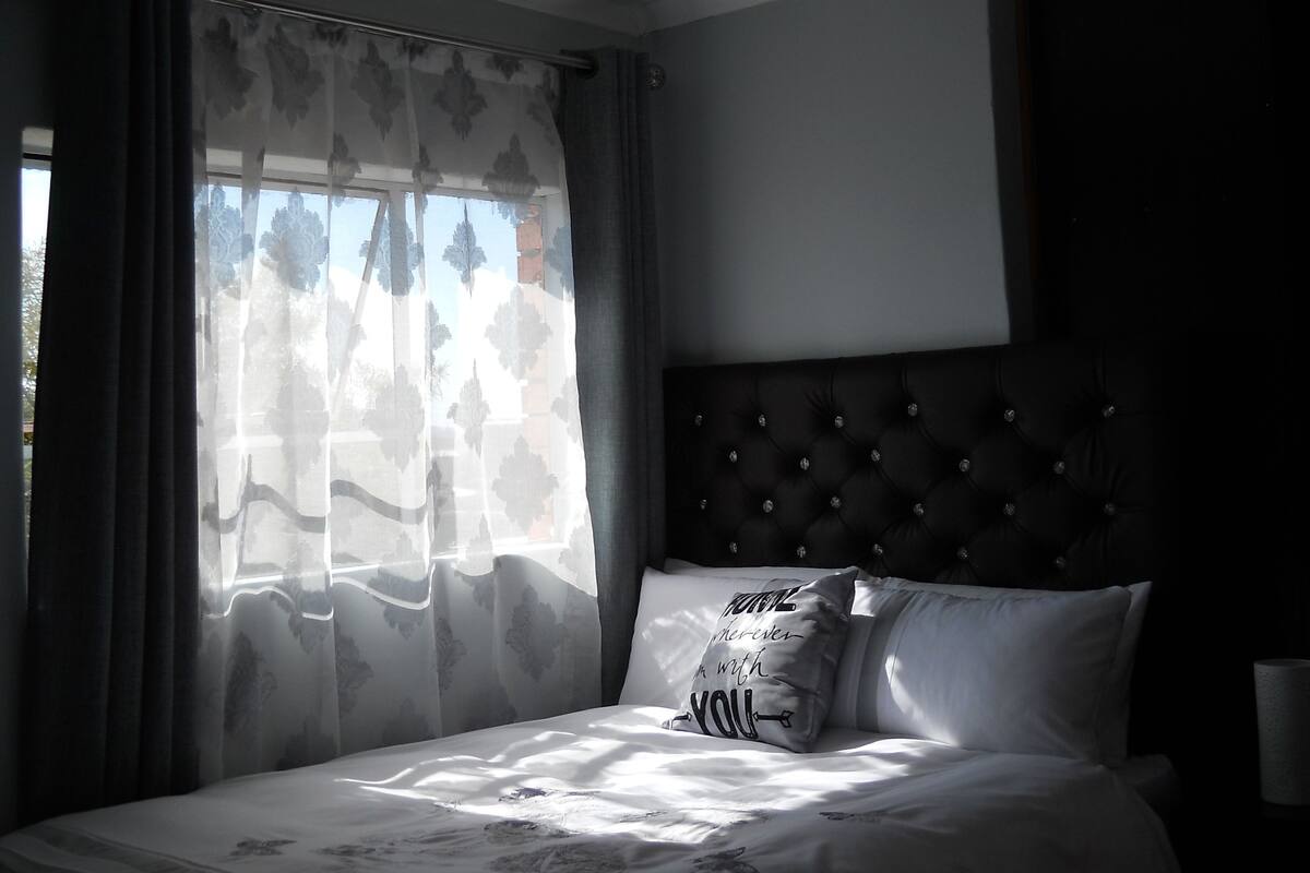 A bedroom with a bed, a tufted headboard, sheer curtains, and a pillow with a quote.