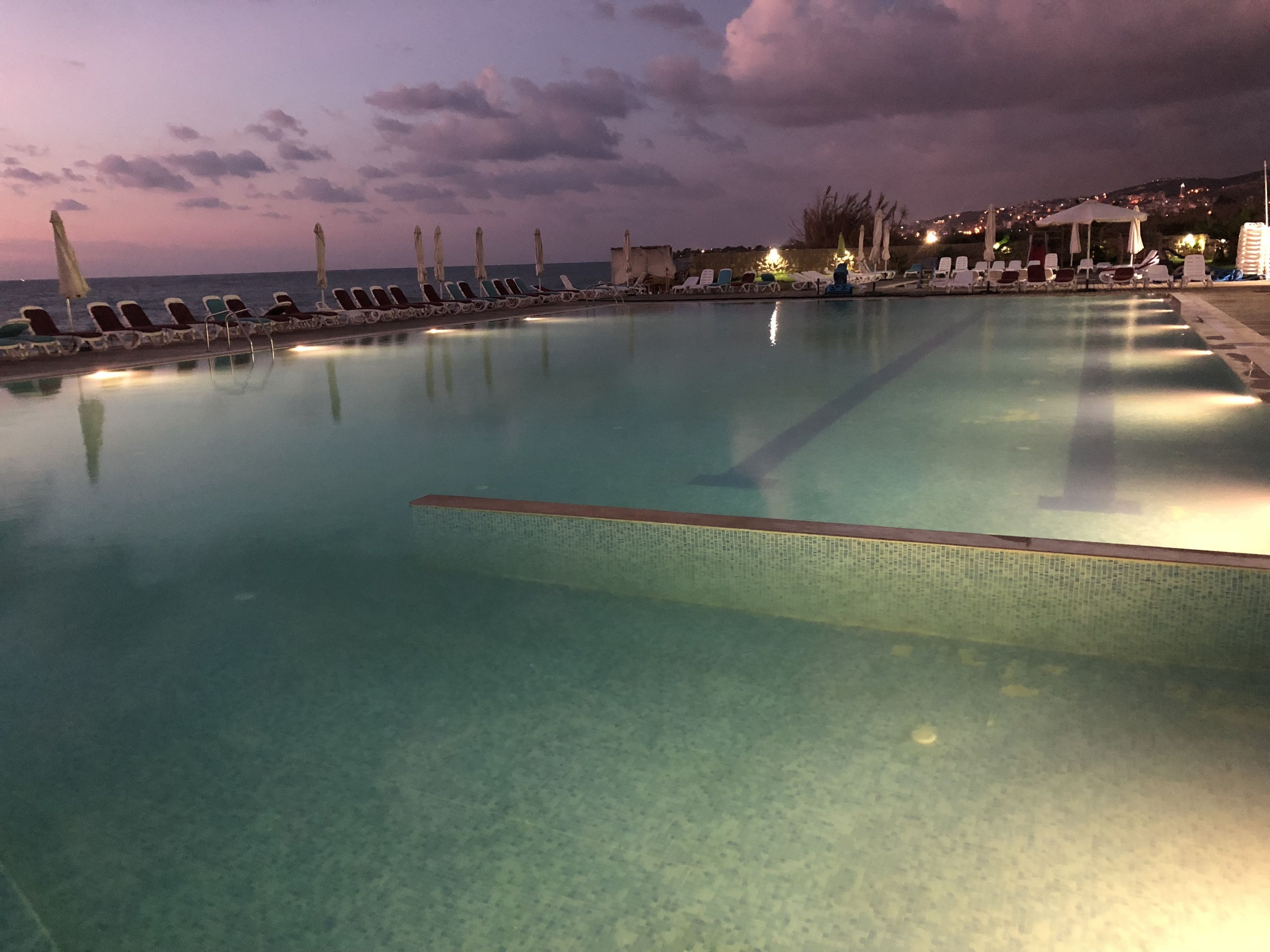 A swimming pool with lounge chairs and umbrellas, surrounded by a poolside area with a view of the sea and distant buildings.
